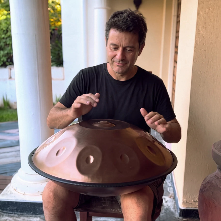 A man playing a bronze handpan on his lap, seated on a shaded veranda