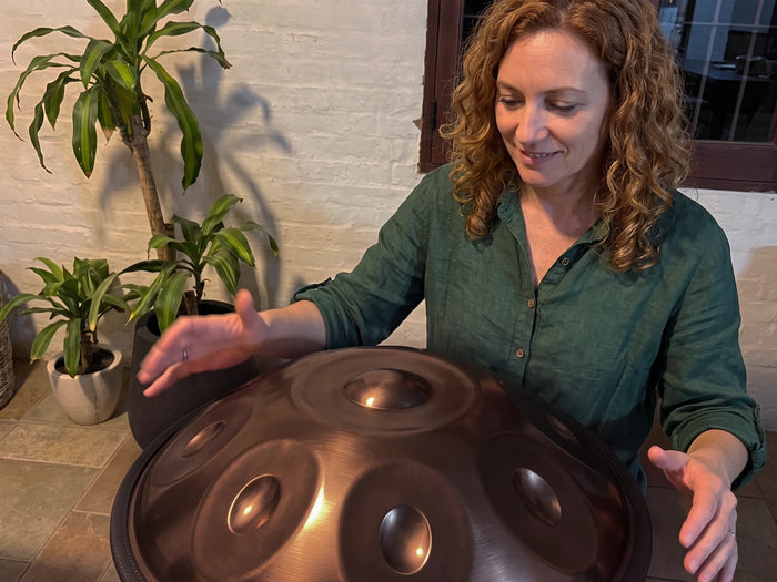 A woman playing a bronze handpan at home, one hand above the tone fields, with plants and a white brick wall behind her