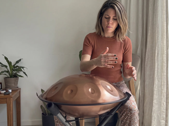 A woman playing a bronze handpan on its stand at home, both hands above the tone fields, with a plant and a linen curtain behind her
