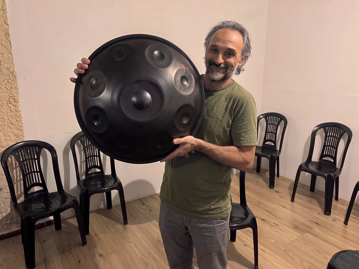 A man holding a large black stainless steel handpan in a rehearsal room, with the tone fields facing the camera and chairs along the white wall behind him