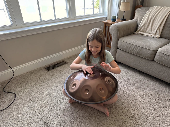 A child sitting on a living room floor, playing a steel handpan with a bronze finish on her lap