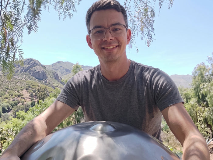 A man holding a brushed stainless steel handpan outdoors, with green hills and mountains behind him under a clear sky